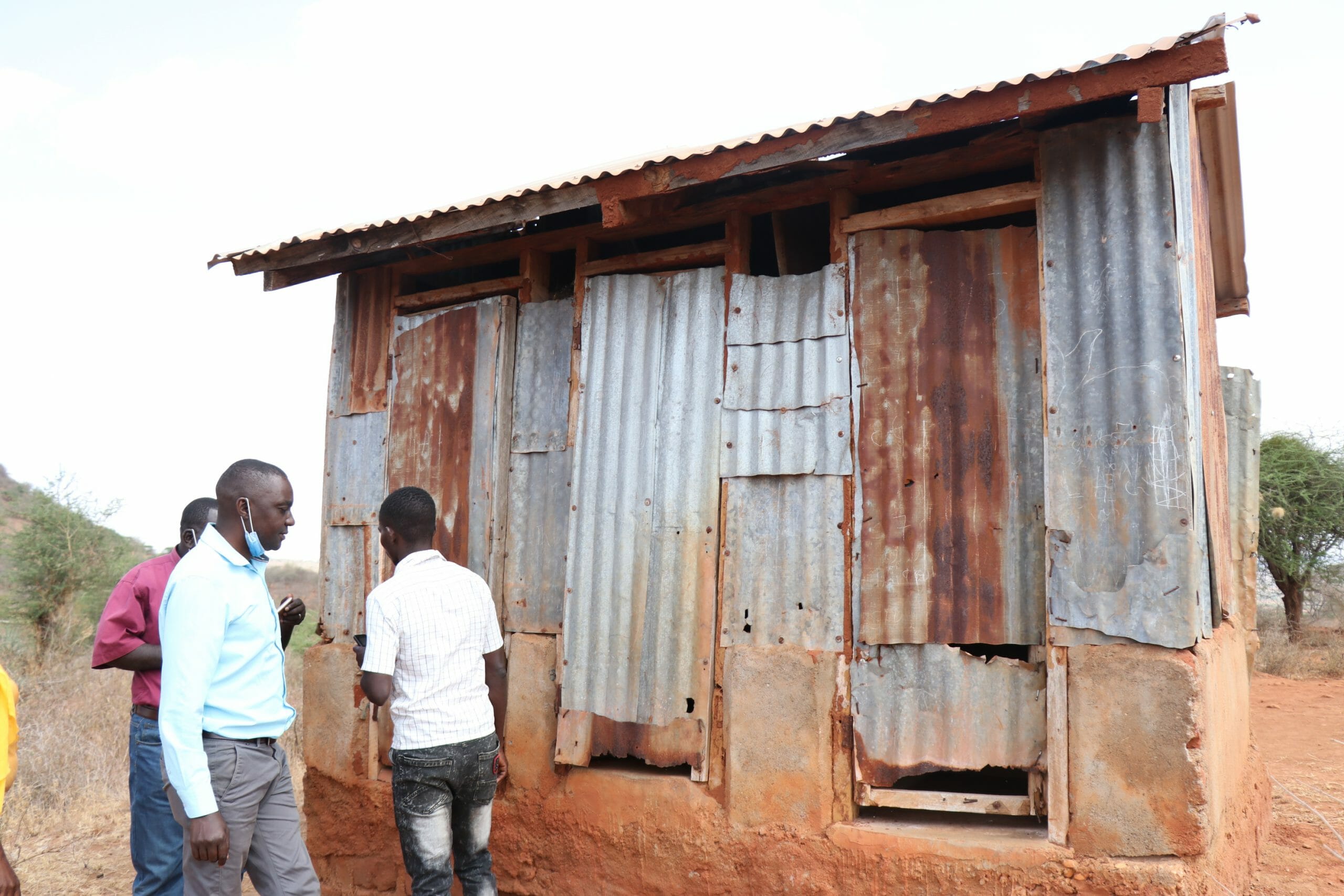 The old toilet block with iron sheet walls and doors with 3 men in front